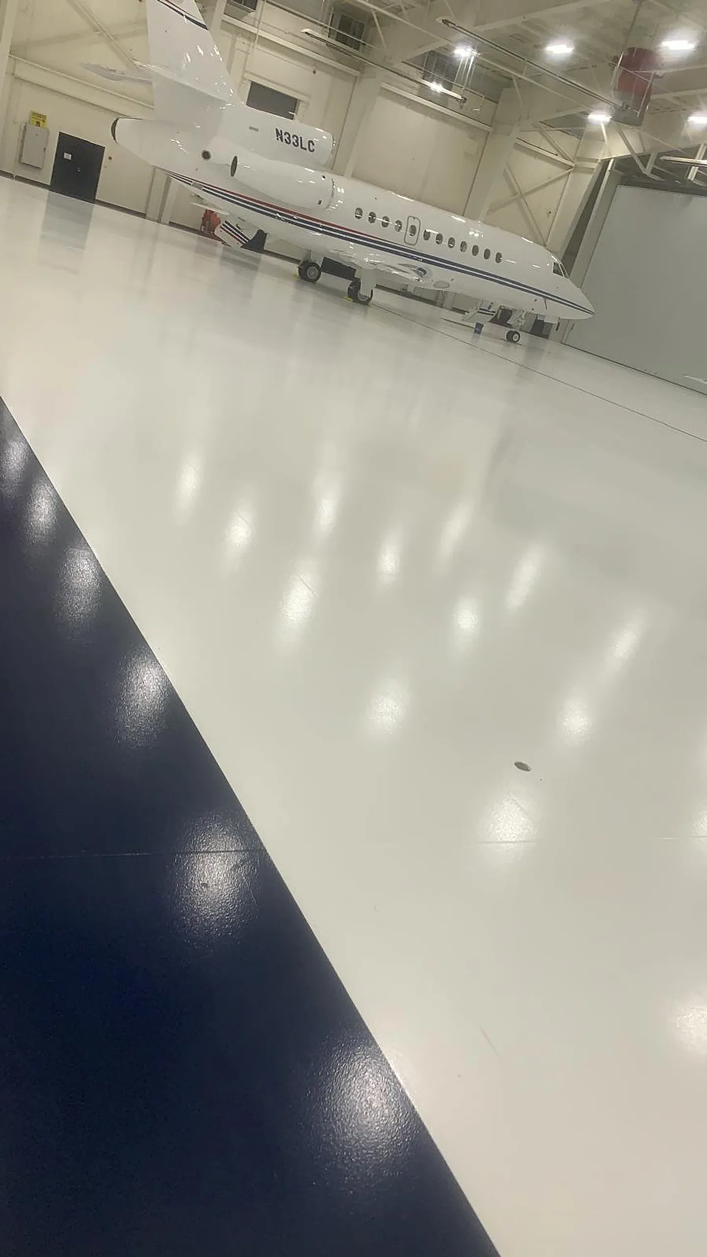 Glossy white resinous floor coating with a navy accent stripe in an aircraft hangar, with a private jet parked in the background — real SFS project