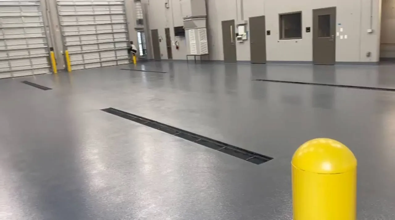 Grey broadcast-coated floor in a vehicle maintenance bay with trench drains, yellow bollards and overhead coiling doors