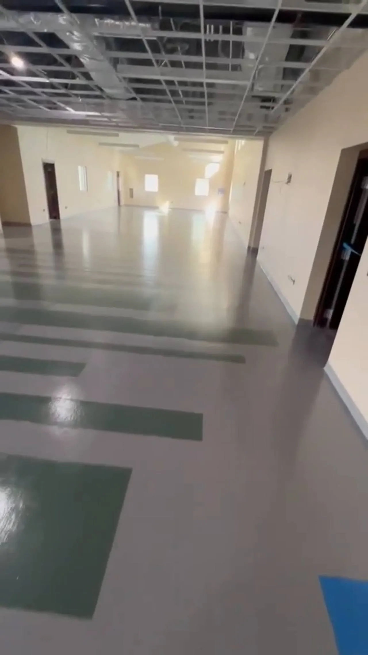 Large institutional room with tan block walls, an exposed ceiling grid, and a resinous floor finished in broad green and grey colour bands